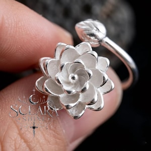 May include: A silver ring featuring a detailed succulent flower design. The ring's focal point is a fully bloomed flower with layered petals, and a bud on the band. The ring is held by a person.