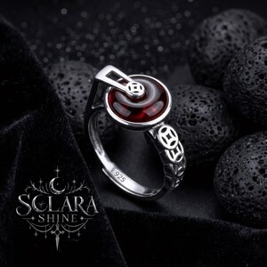 May include: A silver ring with a round, deep red stone center. The ring features an open design with decorative cutouts and a small, rectangular accent above the stone. The ring is set against a dark background.