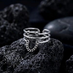 May include: A silver-toned ring with a layered design. The ring features three bands, a small chain, and a rectangular black accent. The ring is displayed on a dark, textured surface.