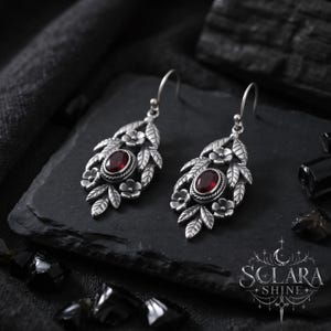 Può includere: Un paio di orecchini pendenti in argento con una pietra preziosa rosso scuro dal taglio ovale al centro, circondata da foglie in metallo dettagliate e piccoli fiori. Gli orecchini pendono da ganci in argento e includono il testo SCLARA SHINE.