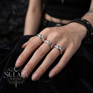 May include: Three silver rings with gemstone details are shown on a hand. The rings feature blue, yellow, and dark-coloured stones. The rings are set against a dark background. The brand name "SCLARA SHINE" is visible in the lower left corner.