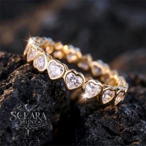 Peut inclure: Une bague dorée ornée de pierres transparentes en forme de cœur. La bague est posée sur une surface sombre et texturée. Le design de la bague présente un motif continu de pierres en forme de cœur, créant un effet scintillant. C'est un bijou.