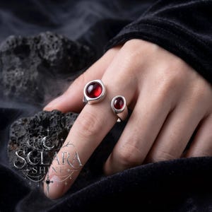 May include: Two silver rings with oval, deep red gemstones. The larger ring is on the index finger, and the smaller ring is on the pinky finger. The rings have a minimalist design, with a smooth, polished finish. The rings are set against a dark background.