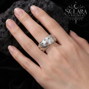 May include: A wide silver band ring with a textured, matte finish featuring multiple small, colorful embedded gemstones in various hues including blue, green, and orange, each set within a small gold-toned circular bezel setting.