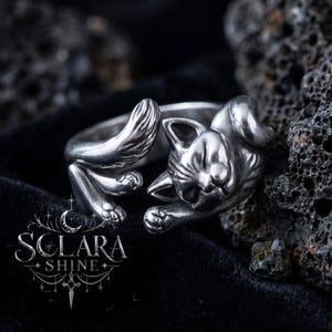 May include: A silver-colored ring in the shape of a sleeping cat. The ring features the cat's head, paws, and tail, with intricate detailing. The ring is displayed on a black surface. The brand name "SOLARA SHINE" is visible.