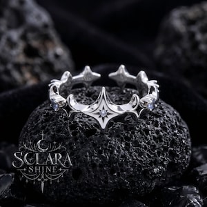 Puede incluir: Un anillo de plata con un diseño de estrella y media luna. El anillo presenta un diseño central de estrella con una pequeña gema y está rodeado de detalles en forma de estrella. El anillo está marcado con "S925" y la marca "Sclara Shine" es visible.