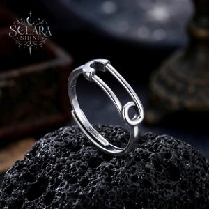 Könnte beinhalten: Ein silberner Ring in Form einer Sicherheitsnadel, vor einem dunklen, strukturierten Hintergrund. Der verstellbare Ring trägt die Markierung "S925". Ein einzigartiges Schmuckstück.