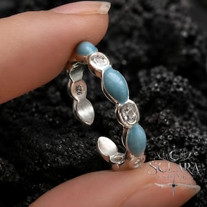 Puede incluir: Un anillo plateado con esmalte turquesa ovalado y piedras redondas y transparentes. El anillo se sostiene entre dos dedos sobre un fondo oscuro y texturizado. El anillo está marcado con "925".