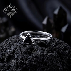 May include: A silver ring with a black triangular stone. The ring is set on a dark, textured surface. The ring's band is thin and polished, and the triangular stone is centered on the ring.
