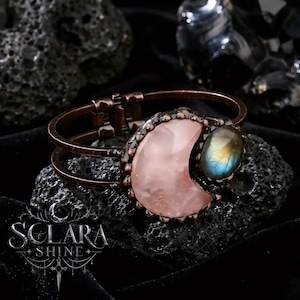 May include: A copper-toned metal cuff bracelet featuring a large, smooth, pale pink crescent-shaped stone and a smaller, oval-shaped iridescent blue and gold labradorite stone. The bracelet has a hinged opening and a dark, weathered metal finish.