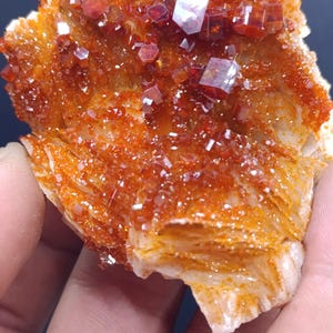 Puede incluir: Un racimo de cristales de vanadinita de color naranja vibrante y rojo intenso con estructura hexagonal. El espécimen mineral presenta una disposición densa de pequeñas formaciones cristalinas translúcidas y reflectantes sobre una base pálida y texturizada.