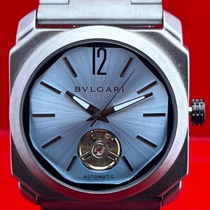May include: A silver-toned stainless steel wristwatch with a brushed metal finish, featuring a light blue sunray dial, a prominent numeral 12, silver baton indices, a visible balance wheel at the six o'clock position, and the text BVLGARI AUTOMATIC.