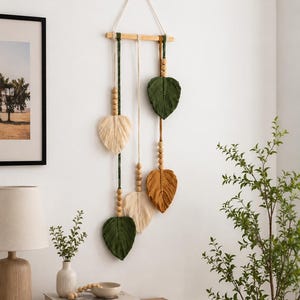 May include: A macrame wall hanging featuring a horizontal wooden dowel suspended by a cord. Five vertical strands hang from the dowel, each decorated with natural wooden beads and finished with a fiber leaf shape in cream, olive, or mustard.
