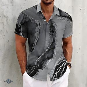 May include: Short-sleeved shirt with a grey and black abstract marble pattern. The shirt has a classic collar and a single button closure. The design incorporates white and silver details, giving a textured look. The shirt is worn with white trousers.