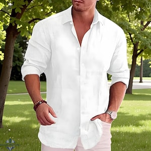 May include: A white, long-sleeved button-down shirt. The shirt is unbuttoned at the top and has a relaxed fit. The person is wearing a watch and a bracelet. The background is a green, grassy area with trees.