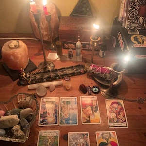 Peut inclure: Une surface de table en bois avec sept cartes de tarot dispos&eacute;es sur deux rang&eacute;es, incluant le Roi d'&Eacute;p&eacute;es et le Deux de Coupes. La sc&egrave;ne comprend des bougies allum&eacute;es dans des bougeoirs, une lampe &agrave; sel, des cristaux et un bol de pierres.