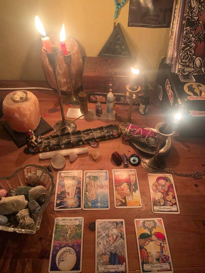 Peut inclure: Une surface de table en bois avec sept cartes de tarot dispos&eacute;es sur deux rang&eacute;es, incluant le Roi d'&Eacute;p&eacute;es et le Deux de Coupes. La sc&egrave;ne comprend des bougies allum&eacute;es dans des bougeoirs, une lampe &agrave; sel, des cristaux et un bol de pierres.