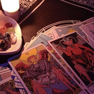 Peut inclure: Un ensemble de cartes de tarot repr&eacute;sentant L'Amoureux et Le Pendu, accompagn&eacute; d'une coupelle blanche contenant divers petits cristaux polis et une bougie pilier blanche allum&eacute;e, dispos&eacute;s sur un tissu noir &agrave; bordure d&eacute;corative blanche.