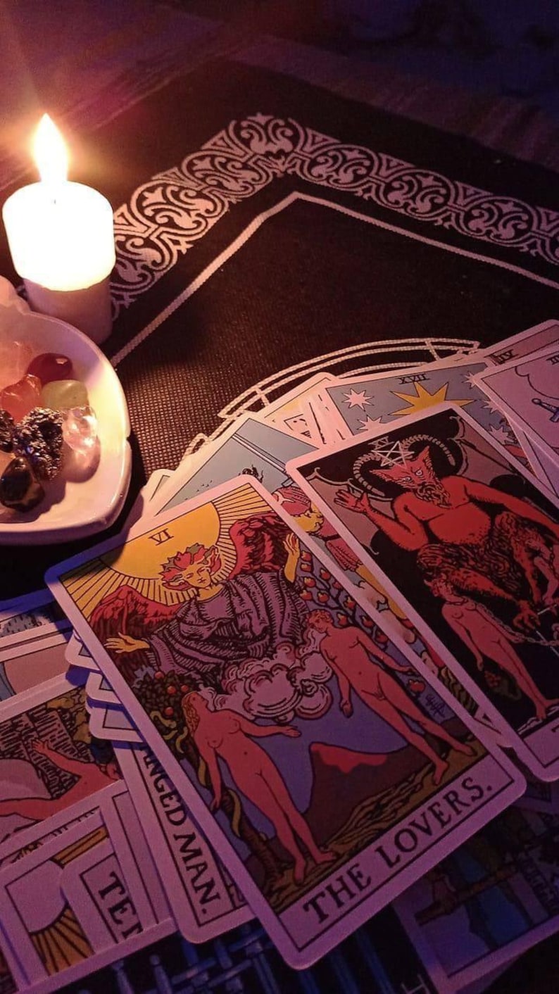 Peut inclure: Un ensemble de cartes de tarot repr&eacute;sentant L'Amoureux et Le Pendu, accompagn&eacute; d'une coupelle blanche contenant divers petits cristaux polis et une bougie pilier blanche allum&eacute;e, dispos&eacute;s sur un tissu noir &agrave; bordure d&eacute;corative blanche.
