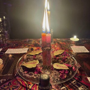 Peut inclure: Deux bougies hautes et fines, une orange et une rose, sont li&eacute;es ensemble avec de la ficelle et allum&eacute;es, au centre d'une assiette en verre transparent. L'assiette est remplie de p&eacute;tales de rose s&eacute;ch&eacute;s, d'herbes et de trois feuilles de laurier.