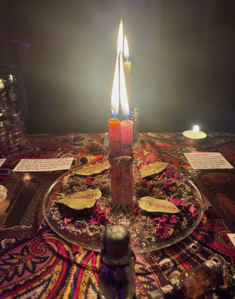 Peut inclure: Deux bougies hautes et fines, une orange et une rose, sont li&eacute;es ensemble avec de la ficelle et allum&eacute;es, au centre d'une assiette en verre transparent. L'assiette est remplie de p&eacute;tales de rose s&eacute;ch&eacute;s, d'herbes et de trois feuilles de laurier.