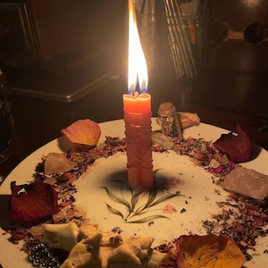 Peut inclure: Une bougie double orange allum&eacute;e et entour&eacute;e de fil m&eacute;tallique fin, plac&eacute;e au centre d'une assiette blanche &agrave; motif floral. L'assiette est garnie de p&eacute;tales de rose s&eacute;ch&eacute;s, d'herbes &eacute;parpill&eacute;es, de petits cristaux, d'un flacon et d'un coquillage.
