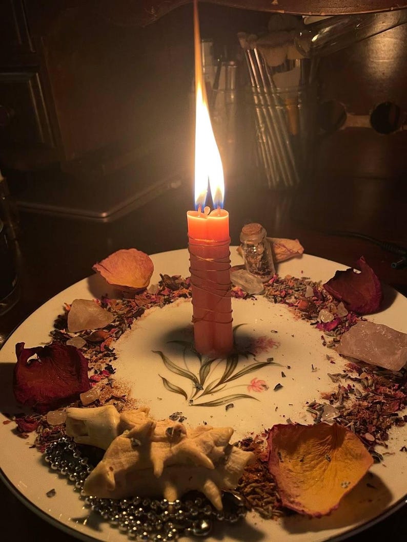 Peut inclure: Une bougie double orange allum&eacute;e et entour&eacute;e de fil m&eacute;tallique fin, plac&eacute;e au centre d'une assiette blanche &agrave; motif floral. L'assiette est garnie de p&eacute;tales de rose s&eacute;ch&eacute;s, d'herbes &eacute;parpill&eacute;es, de petits cristaux, d'un flacon et d'un coquillage.