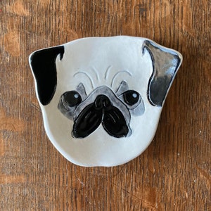 May include: A white ceramic dish shaped like a pug's face. The dish has black and gray painted details, including the pug's eyes, nose, and ears.