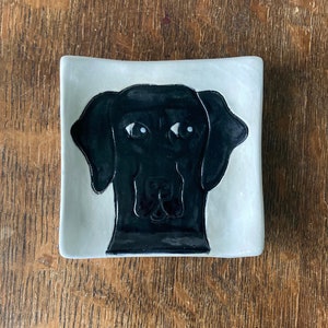 May include: A white ceramic dish with a black dog silhouette. The dog has white eyes and a black nose.