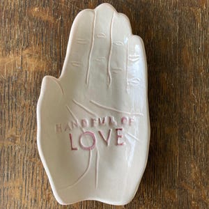 May include: A cream-colored ceramic hand-shaped dish with the words "HANDFUL OF LOVE" in pink lettering. The dish has detailed finger lines and is set against a wooden background.