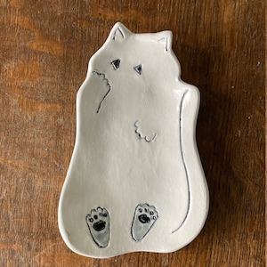 May include: White ceramic cat dish with black paw prints and a simple outline of a cat. The dish is shaped like a cat sitting on its hind legs.