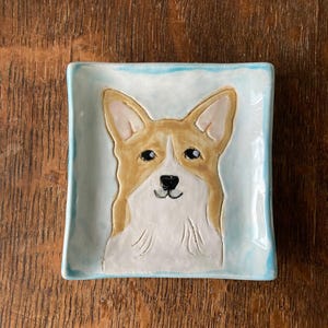 May include: Square ceramic dish with a light blue border and a corgi dog design. The corgi has tan and white fur, black eyes, and a black nose. The dish is on a wooden surface.