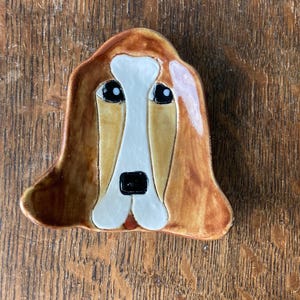 May include: A ceramic dish shaped like a basset hound's face. The dish is painted in shades of brown, white, and black. The dog's eyes are black with white pupils, and its nose is black. The dish is a unique and whimsical piece of pottery.