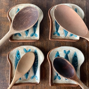 Op de afbeelding: Vier keramische lepelsteunen in de vorm van sneetjes brood met blauwe visontwerpen. Elke steun houdt een houten lepel.