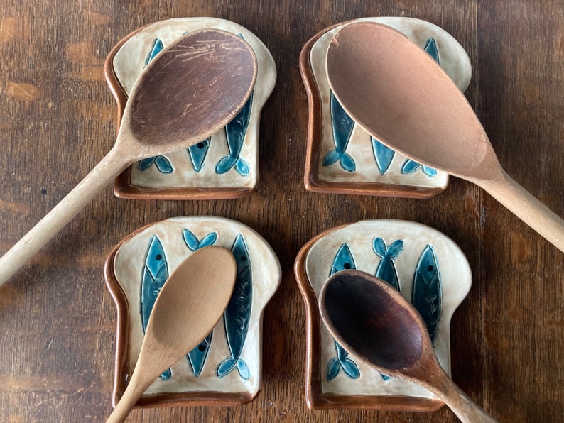 Op de afbeelding: Vier keramische lepelsteunen in de vorm van sneetjes brood met blauwe visontwerpen. Elke steun houdt een houten lepel.