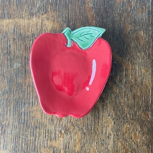 May include: A small, glossy ceramic dish shaped like a red apple with a single green leaf attached at the top. The surface of the apple features a smooth, reflective finish with a white highlight, creating a bright and polished appearance.
