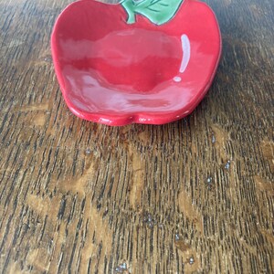 May include: A small, glossy ceramic dish shaped like an apple. The dish is bright red with a smooth, reflective surface and features a single, raised green leaf detail at the top center. The bowl has a shallow, rounded interior basin.