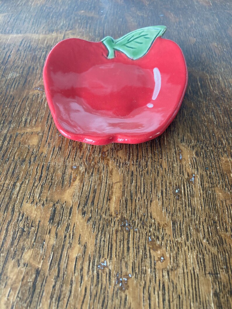 May include: A small, glossy ceramic dish shaped like an apple. The dish is bright red with a smooth, reflective surface and features a single, raised green leaf detail at the top center. The bowl has a shallow, rounded interior basin.