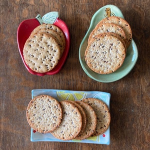 May include: Three ceramic dishes holding round, seeded crackers. One dish is red and apple-shaped, one is light green and pear-shaped, and one is a rectangular tray with a yellow and blue lemon slice pattern. Each dish contains a stack of crackers.