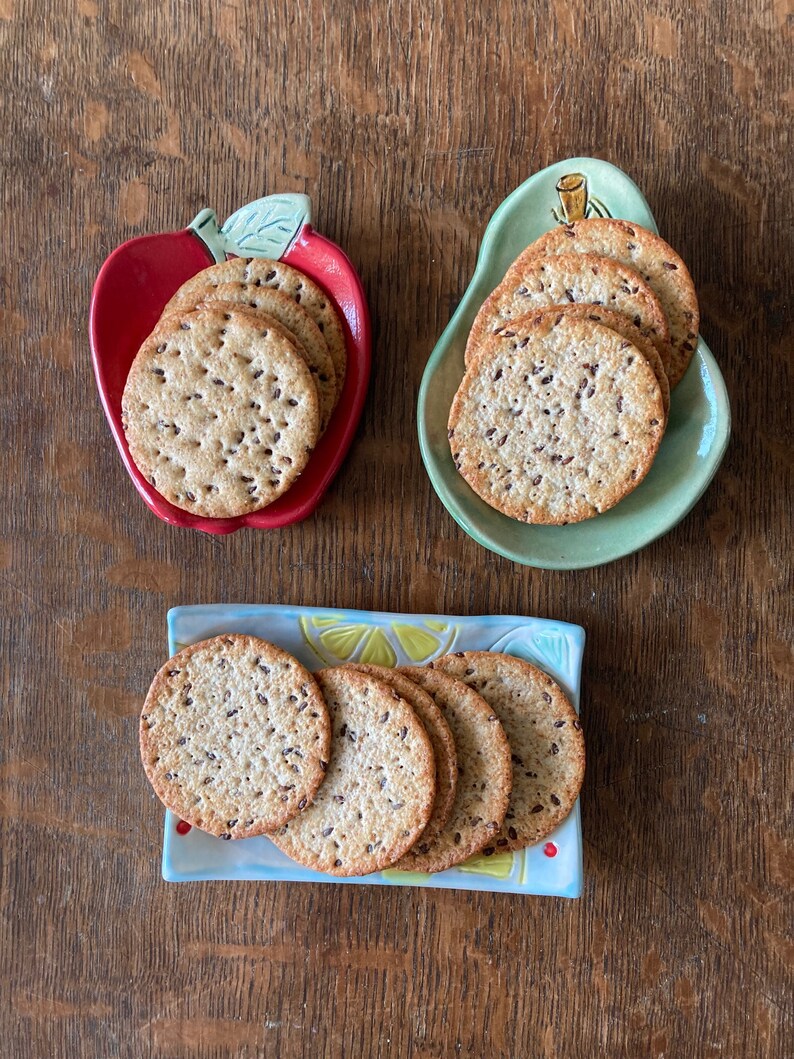May include: Three ceramic dishes holding round, seeded crackers. One dish is red and apple-shaped, one is light green and pear-shaped, and one is a rectangular tray with a yellow and blue lemon slice pattern. Each dish contains a stack of crackers.