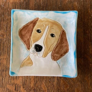 May include: A ceramic dish with a blue border and a white background features a hand-painted image of a beagle dog with brown and white fur. The dog's head is tilted to the side, and its eyes are looking directly at the viewer.