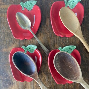 May include: Four ceramic apple-shaped spoon rests in a glossy red finish with green leaves. Three wooden spoons and one silver-toned metal spoon are placed on top of the spoon rests. The apple-shaped dishes feature a white highlight detail.
