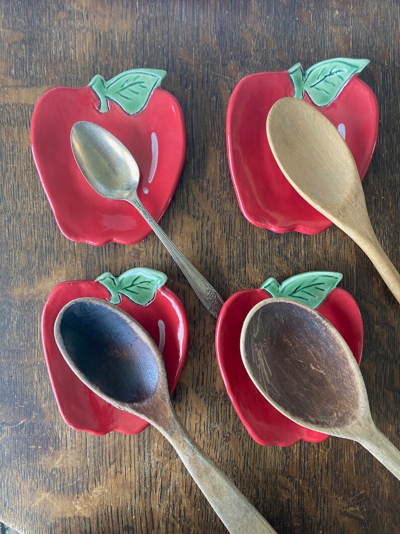 May include: Four ceramic apple-shaped spoon rests in a glossy red finish with green leaves. Three wooden spoons and one silver-toned metal spoon are placed on top of the spoon rests. The apple-shaped dishes feature a white highlight detail.