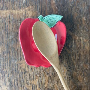 May include: A red ceramic spoon rest shaped like an apple with a green leaf detail at the top. A light-colored wooden spoon is positioned diagonally across the center of the apple-shaped dish, partially covering the glossy red surface.