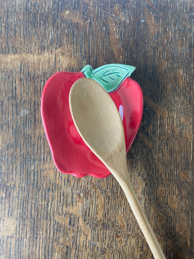 May include: A red ceramic spoon rest shaped like an apple with a green leaf detail at the top. A light-colored wooden spoon is positioned diagonally across the center of the apple-shaped dish, partially covering the glossy red surface.