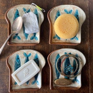 Op de afbeelding: Vier keramische broodvormige schalen met blauwe visontwerpen. Elke schaal bevat een ander item: een zilveren lepel, een theezakje, een gele spons en een set gouden armbanden.