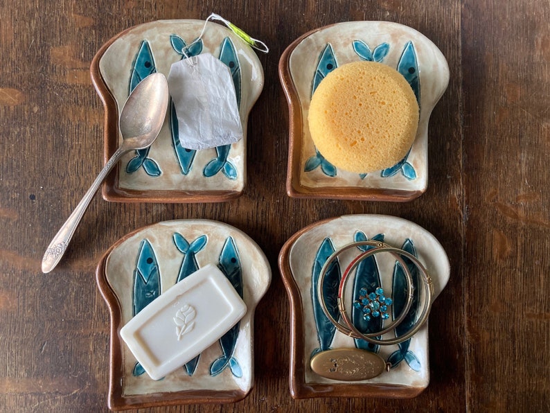 Op de afbeelding: Vier keramische broodvormige schalen met blauwe visontwerpen. Elke schaal bevat een ander item: een zilveren lepel, een theezakje, een gele spons en een set gouden armbanden.