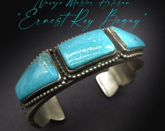 May include: A silver cuff bracelet with three turquoise stones set in the center. The bracelet is decorated with a beaded border. "Navajo Master Artisan Ernest Roy Begay" is written in cursive above the bracelet.