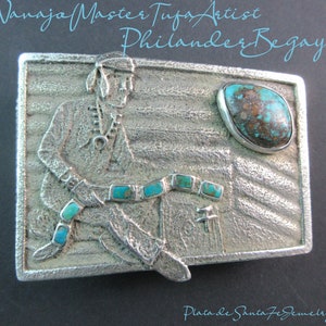 May include: A silver belt buckle with a turquoise inlay depicting a Native American man sitting with a turquoise belt. The buckle is signed "Philander Begay" and "Plata de Santa Fe Jewelry".