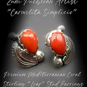 May include: A pair of sterling silver stud earrings featuring a central smooth, bright orange Mediterranean coral stone. Each earring is accented with a metallic leaf motif and small silver spheres. Text reads: Zuni Puebloan Artist Carmelita Simplicio, Premium Mediterranean Coral Sterling Leaf Stud Earrings.